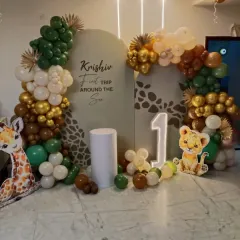 For a child's first birthday, I can create a memorable "First Trip Around the Sun" jungle theme. This setup includes a custom-printed backdrop with a giraffe pattern, surrounded by an organic balloon garland in earthy tones of green, brown, and gold. I also provide character cutouts like a baby giraffe and lion, a light-up marquee number '1', and a pedestal for the cake.