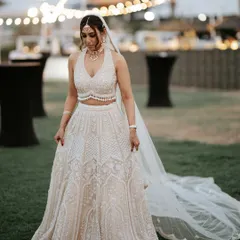 A full-length portrait of the bride in her stunning white lehenga. She looks elegant and serene against the backdrop of the evening reception setup. A moment of quiet grace.
