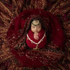 A breathtaking portrait of a newborn girl swaddled in red and surrounded by the rich, intricate embroidery of her mother's wedding lehenga. She wears a tiny, custom made maang tikka to complete the look.