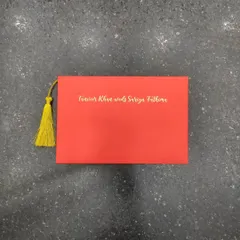 The front of the bold red envelope, featuring the couple's names in elegant gold foil script and a bright yellow tassel for a striking color contrast.