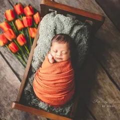 Blooming with love. This newborn is nestled in a tiny wooden bed, swaddled in a warm orange wrap next to fresh tulips, creating a beautiful, artistic portrait that feels like a dream.