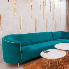 A close-up of the custom-made teal sofa with quilted details and gold piping. This piece adds a luxurious feel and a vibrant splash of color to the living room, set against a marble-finish wall with gold inserts.