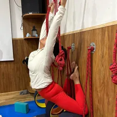 A student in a vibrant red outfit practices a supported backbend. The ropes and wheel provide the necessary lift and stability to work on spinal flexibility without compression.