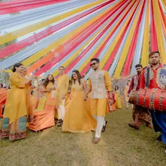 The groom's energetic entry with a dhol player, surrounded by family and friends dressed in yellow. The multi-coloured fabric canopy we installed provides a festive backdrop for this joyous procession.