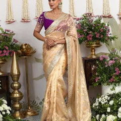 The ethereal beauty of this ivory organza saree is elevated by a contrasting deep purple embroidered blouse. It is a testament to understated luxury, perfect for intimate celebrations where grace and sophistication are key.