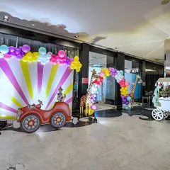 An exciting indoor carnival setup featuring a vibrant photo booth with a circus car cutout. The scene is brought to life with a colorful balloon arch, life-sized animal props like a zebra and giraffe, and a multi-activity cart.