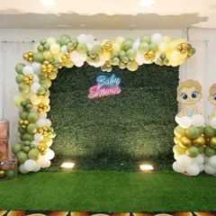 A simple and elegant baby shower photo booth. This design uses a green hedge wall as the backdrop, a 'Baby Shower' neon sign, and a balloon arch in muted green, white, and gold tones.