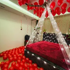 A classic romantic room decoration featuring a bed canopy with fairy lights, red balloons covering the floor and ceiling, and a heart made of rose petals on the bed.