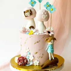 An enchanting woodland-themed cake. This pink cake is decorated with gold leaf, flowers, and adorable toppers including a girl figurine and whimsical mushrooms.