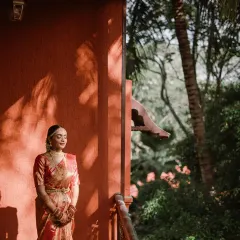 A South Indian bride, bathed in the golden hour light, stands on a balcony. The play of light and shadow on the terracotta wall creates a stunning, artistic portrait that highlights her traditional beauty.