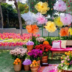 A whimsical and colorful photo booth for a Mehendi party. The backdrop features giant, handcrafted paper flowers, while the foreground is filled with baskets of vibrant chrysanthemums and fresh oranges.