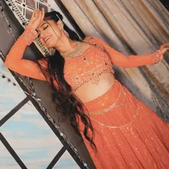 A dramatic, expressive pose from the 'Jhanjhariya' shoot. The vibrant orange lehenga and intricate jewelry create a stunning visual against the artistic backdrop.