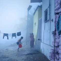 A foggy morning on the Yamuna Ghat. A person washes clothes while life continues in the background, all enveloped in a soft, blue mist that creates a painterly effect.