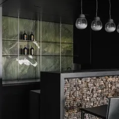 We believe in the honest expression of materials. For this bar area, we conceptualized a Gabion wall, trapping randomly sized stones in a metal wire mesh to create a raw, textural fascia. This Brutalist element is contrasted with a sleek, dark green marble backsplash and minimalist pendant lighting, forming part of a 'black scoop' that unifies the entrance, bar, and kitchen.