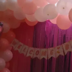 A close-up of the "Welcome Baby" banner from a home decoration setup. The pink shimmer curtains and pastel balloons add a touch of sparkle to the celebration.