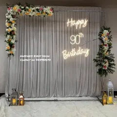 An elegant and simple setup for a 90th birthday. It features a white curtain backdrop, a 'Happy 90th Birthday' neon sign, and minimal floral arrangements with lanterns for a classic look.