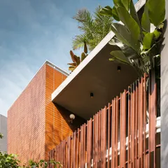 An upward view of the 'Ittige' residence facade, highlighting the interplay between the solid brick volume, the cantilevered concrete roof, and the delicate metal fins. Lush plants spill over from the terrace, reinforcing the connection to nature.