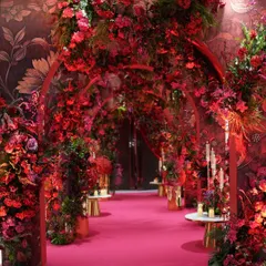 A view down the dramatic red floral entrance tunnel. The path is lined with candles and golden vases, enhancing the warmth and luxury of the design. The floral wallpapered walls add another layer of texture to this immersive experience.