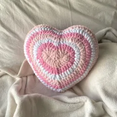 Our crochet heart pillow sitting on a soft, neutral blanket. The y2k-inspired design and colors are fully customizable to match your aesthetic.