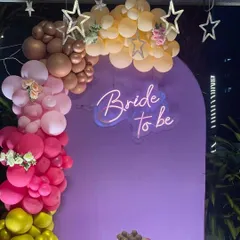 A stunning outdoor setup for a 'Bride to be' party. The purple backdrop with a neon sign is framed by a colorful organic balloon arch in shades of pink, yellow, and brown, with star lights twinkling above.