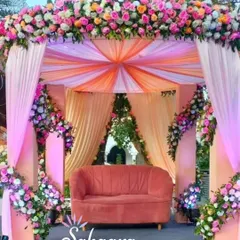 A beautiful outdoor wedding mandap draped in pastel peach and pink fabric. The structure is covered in lush floral arrangements, creating a soft and romantic setting for a daytime wedding.
