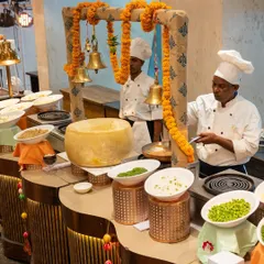 A traditional Indian live cooking station, complete with a large cheese wheel for pasta, surrounded by fresh ingredients. The setup, adorned with marigold garlands and brass bells, adds an authentic and festive touch.