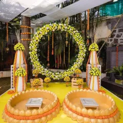 A grand outdoor Haldi setup featuring a large ring of yellow and white flowers. The arrangement includes two decorated urlis, making it perfect for a couple's ceremony.