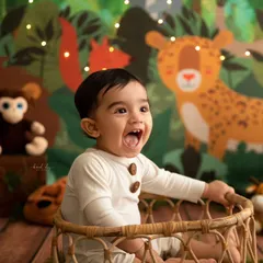 A baby boy laughs with joy in a vibrant jungle-themed setup, surrounded by cartoon animals and fairy lights.