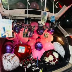 This car trunk decoration includes a "Happy Birthday Baby" cake, a flower bouquet, and a mix of foil and latex balloons.
