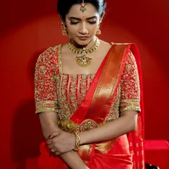 A close-up of a classic South Indian bridal look, featuring a vibrant red Kanjeevaram saree paired with a heavily embroidered blouse. The rich gold zari work and traditional motifs on the blouse showcase the pinnacle of heritage craftsmanship, perfect for a bride seeking authentic grandeur.
