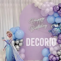 A magical Frozen-themed birthday party featuring a life-sized Elsa cutout. The backdrop is a cool lavender circle with a 'Happy Birthday' neon sign, framed by a balloon garland in shades of purple, blue, silver, and white.