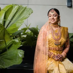A stunning South Indian bride in a golden Kanjeevaram saree, adorned with traditional temple jewelry. Her serene expression and the lush green background create a picture of timeless grace.