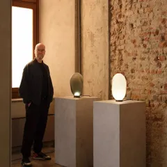 The designer stands beside two Solace table lamps, showcasing the distinct identities of the white and bronze glass versions. This highlights the innovative glassblowing techniques used to create each unique piece.