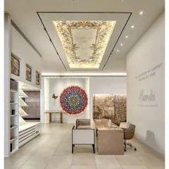The reception area of the Hands Carpets showroom in Mumbai. The design uses a neutral palette, clean lines, and dramatic lighting to frame the carpets as works of art, including a stunning piece displayed on the ceiling.