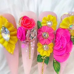 A stunning set of stiletto nails featuring elaborate 3D and 5D floral arrangements in bright pink and yellow, accented with large crystals and gold chains.