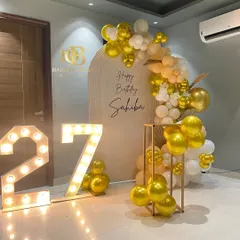 Chic, modern, and personal. This 27th birthday setup features a clean aesthetic with a personalized arched backdrop, a cascade of gold and white balloons, and bold, light-up marquee numbers that create the perfect photo opportunity.