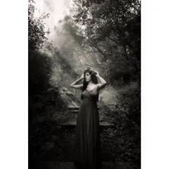 Black and white photo of a model in a dark dress and flower crown, standing in a misty forest with light rays