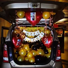 A unique car trunk birthday surprise, perfect for creating a memorable moment. This setup uses gold, black, and red balloons, a "Happy Birthday" banner, and personalized photos attached to heart-shaped foil balloons.