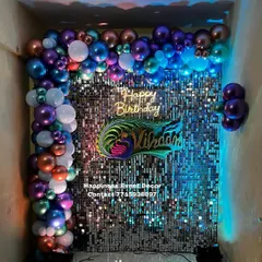 This birthday decor features a stunning silver sequin wall with a multi-colored chrome balloon arch. The custom name prop and 'Happy Birthday' neon sign make it a unique photo backdrop.