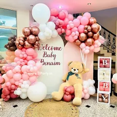 A full view of a chic welcome home party decoration for "Baby Benaim". The setup includes two arched backdrops, an extensive balloon garland in pinks and rose gold, a large teddy bear, and "BABY" letter blocks.