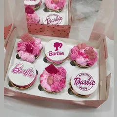A box of custom Barbie-themed cupcakes, perfect for a birthday girl. Each cupcake is 100% eggless, with some featuring detailed edible prints and others topped with pink frosting, sprinkles, and chocolate pieces for a truly magical look.