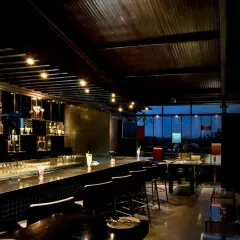 A sophisticated bar lounge designed for curated evenings. The space features a sleek, dark bar, dramatic overhead lighting, and comfortable seating, all set against a backdrop of the city skyline at dusk.