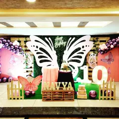 For Navya's butterfly party, the centerpiece is a set of large, elegant butterfly wings against a green wall, perfect for photos. The stage is framed with purple chrome balloon arrangements and includes a personalized name sign and a small picket fence for a whimsical garden touch.