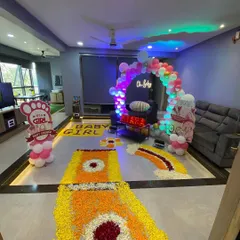 A full view of a living room decorated for a baby's homecoming, with a flower rangoli path, a balloon arch, and a neon 'Oh Baby' sign.