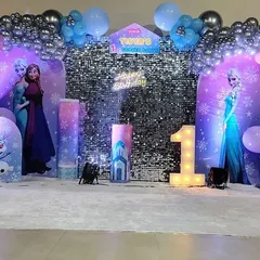 A magical 'Frozen' world for Tisya's 1st birthday celebration. This full-stage setup featured character cutouts of Elsa, Anna, and Olaf against a shimmering silver backdrop, with balloon arches in shades of blue, purple, and silver to create a winter wonderland.