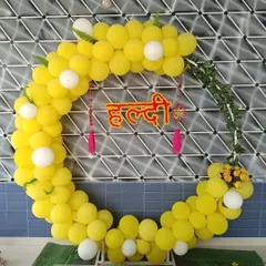 A modern take on Haldi decor, this circular backdrop is made of bright yellow and white balloons. A custom 'Haldi' sign in Hindi script hangs in the center, accented with green vines for a fresh look.