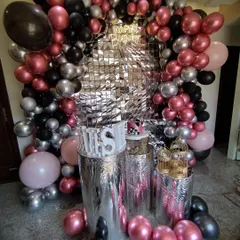 A glamorous party setup with a silver shimmer wall and an organic balloon garland in metallic pink, silver, and black. The plinths are wrapped in silver foil for extra shine.