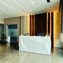 A direct view of the faceted, glossy white reception desk, which serves as the focal point of this Ambala office lobby. The back wall features dark wood panels with vertical accent lighting, adding depth and sophistication.