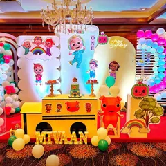 A colorful Cocomelon first birthday party stage. The design includes a large school bus prop, multiple character cutouts, and bright balloon displays, creating a playful atmosphere.
