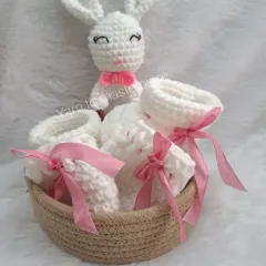 This newborn gift hamper includes soft white booties with pink ribbons and a matching bunny rattle, all presented in a charming woven basket.